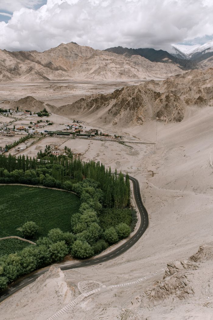 Drone view of narrow road going among green trees and desert with mountains with settlement in distance against cloudy sky in nature