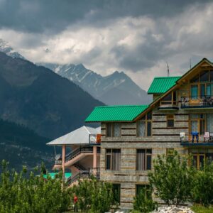 Modern mountain house with green roof, set against snow-capped peaks under cloudy skies.